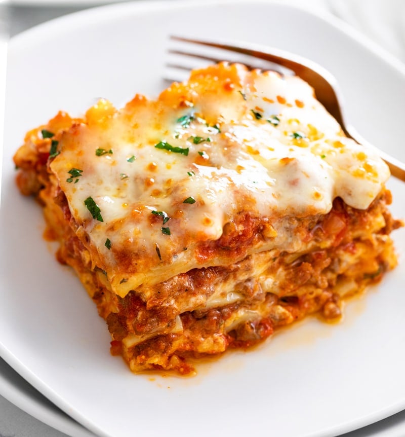A white plate with Lasagna on it with a fork in the background.
